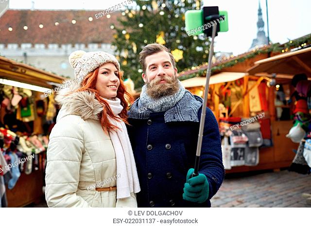holidays, winter, christmas, technology and people concept - happy couple of tourists in warm clothes taking picture with smartphone selfie stick in old town