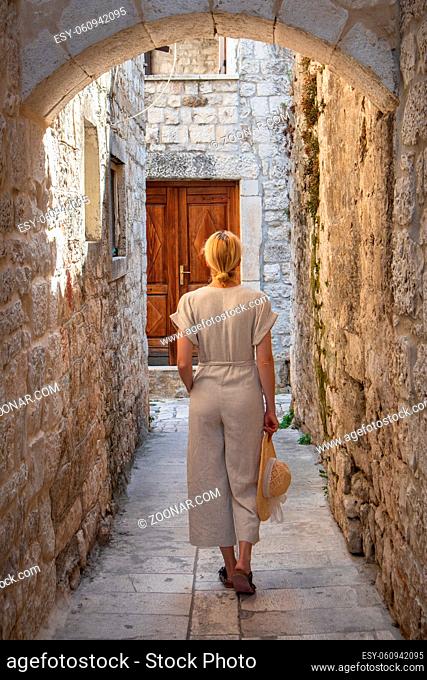 Rear view of beautiful blonde young female traveler wearing straw sun hat sightseeing and enjoying summer vacation in an old traditional costal town at Adriatic...