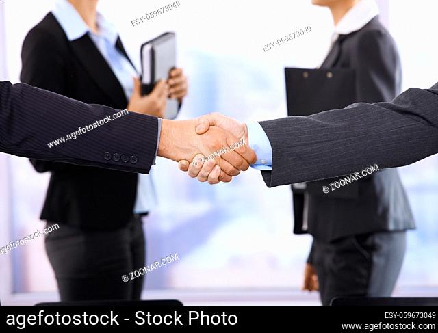Closeup handshake in focus, businesswomen in background in meeting room