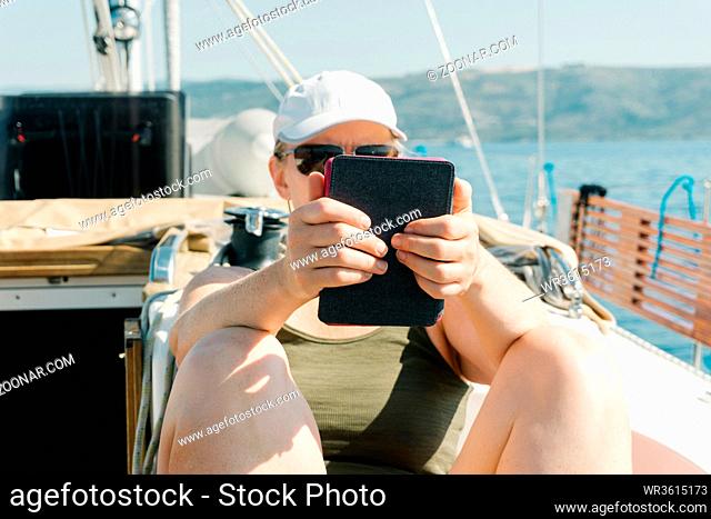 Woman sitting on deck of a yacht enjoying reading in her ebook. Travel and vacation concept
