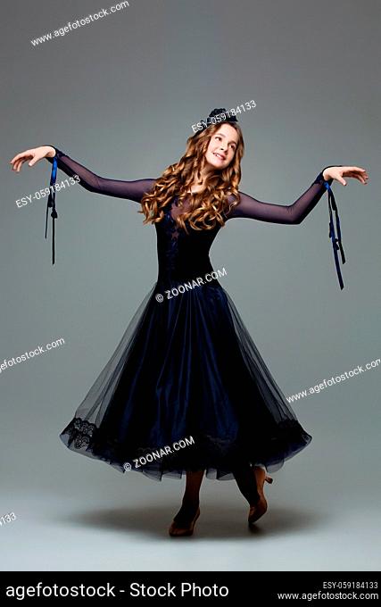 Beautiful teenager ballroom dancer with long blond hair in long dark blue dress making curtsy. Studio shot on grey background. Copy space