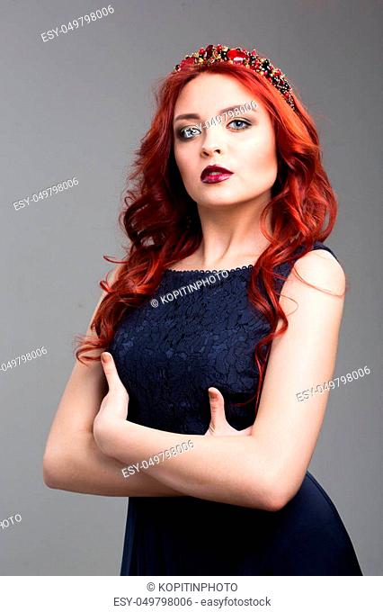 Beautiful red-haired fashion model posing in evening dress and in the diadem over dark background. female gestures of seduction. body language