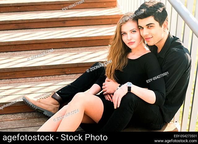 Beautiful young multiracial couple, student couple in love, sit wooden staircase in the city. Beautiful Turkish brunette guy hug a Caucasian girl in black...