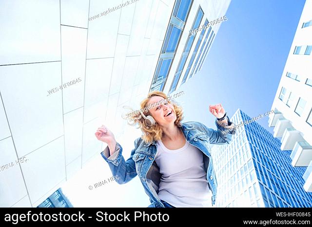 Happy woman enjoying music through wireless headphones standing amidst buildings