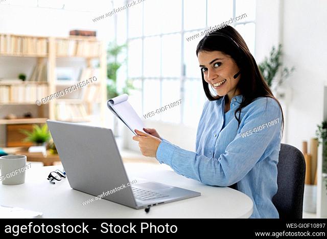 Customer service representative holding note pad while sitting by desk at office