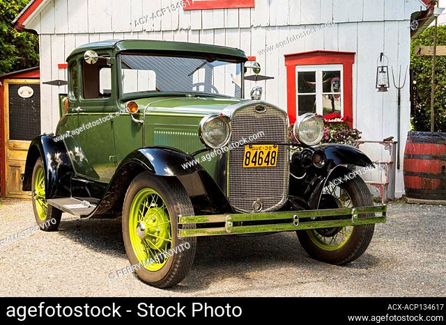 1930 Ford Model A Coupe classic automobile in green with black fenders and tan leather rumble seat in front of old white and red barn-garage in background