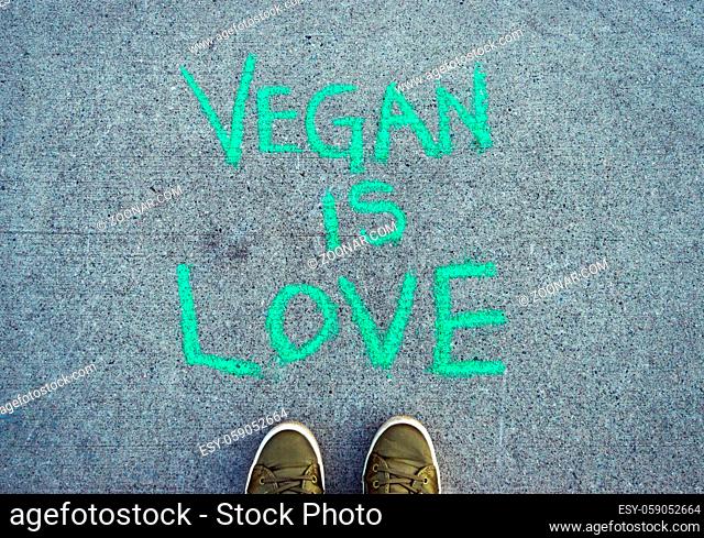 Text words Vegan Is Love in green chalk lettering on cement sidewalk with POV green casual shoes