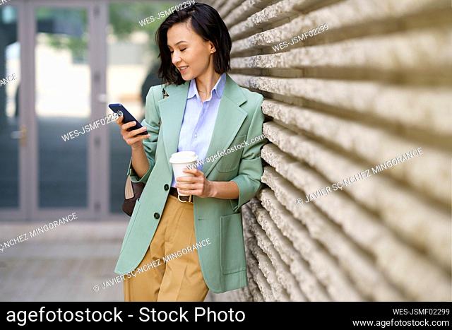 Businesswoman using smart phone while leaning on wall