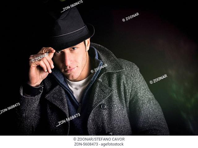 Attractive young man at night, wearing winter coat, tipping fedora hat while looking at camera
