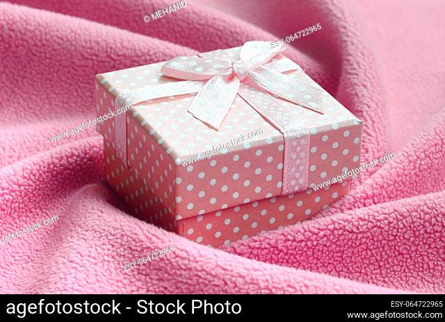 A small gift box in pink with a small bow lies on a blanket of soft and furry light pink fleece fabric with a lot of relief folds