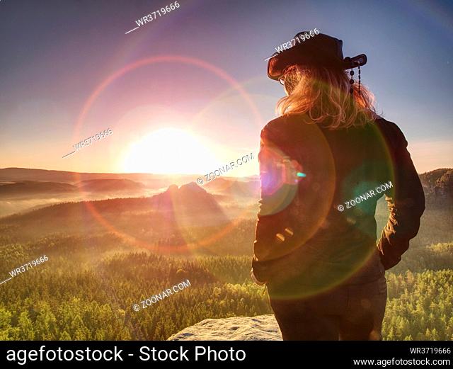 Cheering young woman traveler hiking on rocky mountain promontory above sharp tree tops