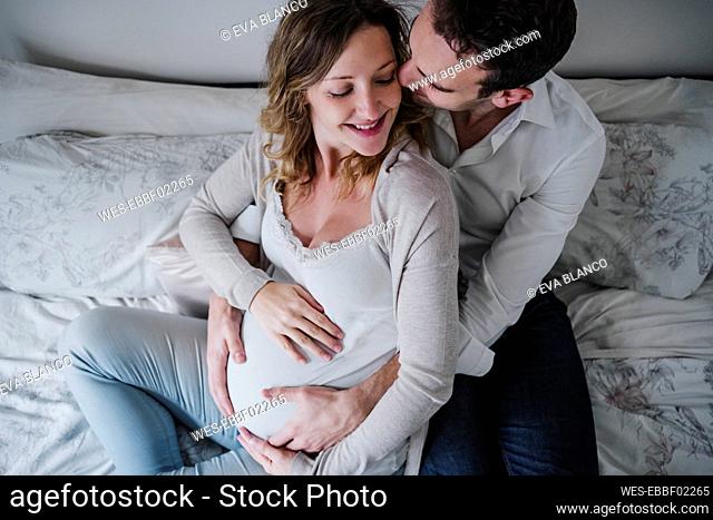 Man with hands on pregnant wife's stomach romancing with her on bed at home