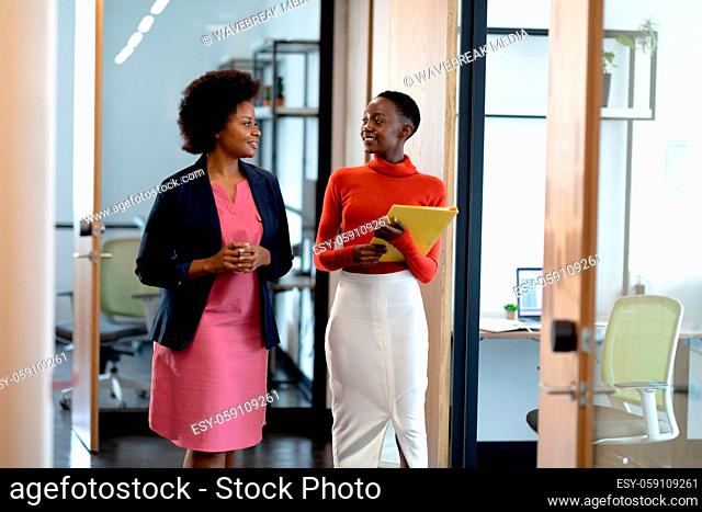 Happy african american female business colleagues walking through corridor holding documents talking