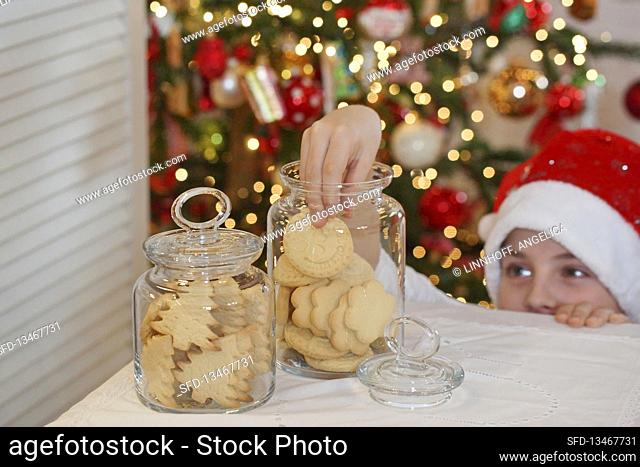 A boy wearing a Santa hat reaching into a jar of biscuits