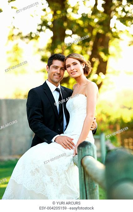 Stunning bride sitting on a hedge with her groom holding his arm around her while they smile at the camera - portrait