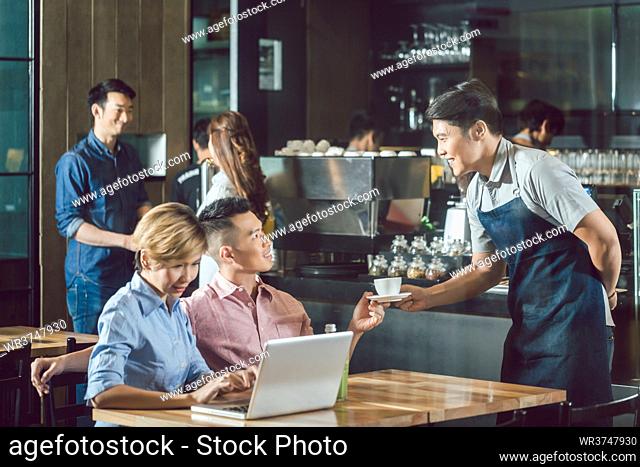 Happy waiter serving coffee to male customer sitting beside woman using laptop