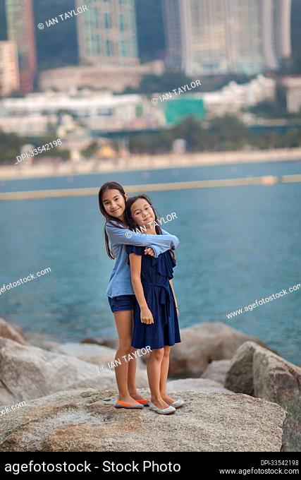 Young sisters stand in an embrace on boulders at Repulse Bay in Hong Kong; Hong Kong, China