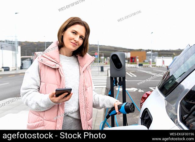 Woman holding charger and smart phone by electric car at station