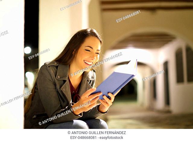 Happy woman reading a paper book sitting in the night in the street