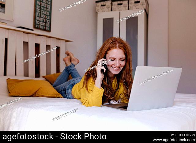 Smiling woman lying on front talking on smart phone in bedroom sitting at home