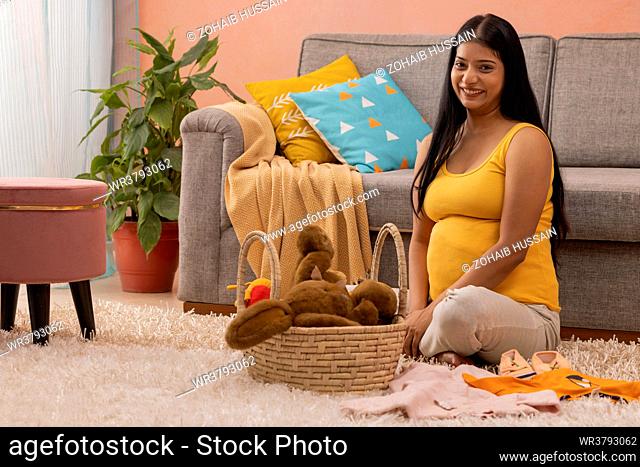 Happy pregnant woman sitting on floor with baby clothes and toys