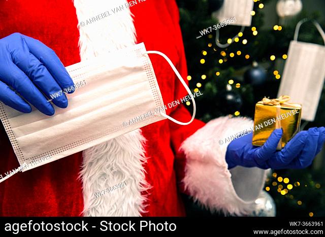 Santa Claus with gloves and face mask for Coronavirus by the Christmas tree, holding a golden present, Covid-19 and Christmas safety concept closeup
