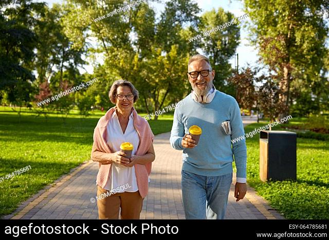 Happy fashioned elderly couple having coffee break walking in park. Romantic aged mature man and woman looking happy enjoying healthy lifestyle on pension