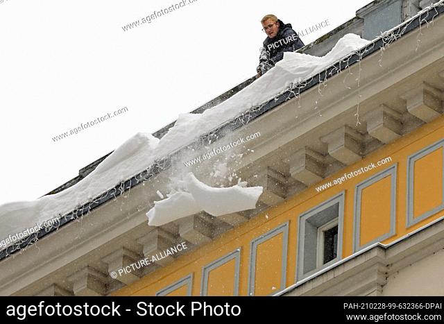 09 February 2021, Saxony-Anhalt, Magdeburg: A man releases overhanging snow slabs from a roof. His colleague secures the footpath