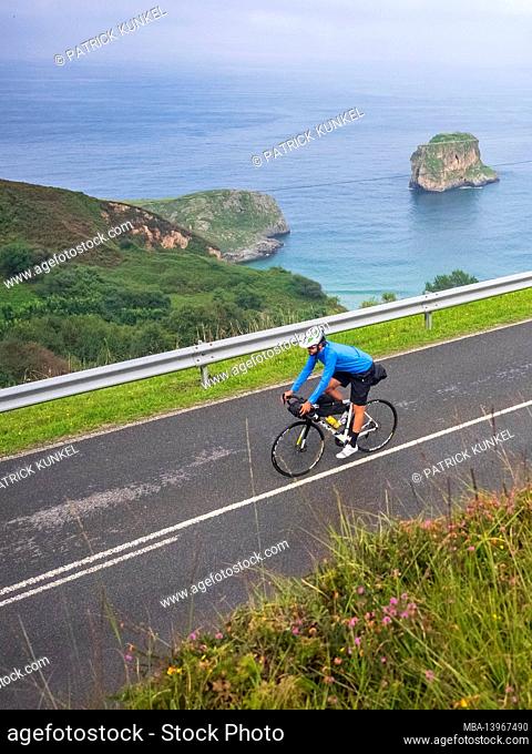 Road bike tour with luggage, bikepacking on a lonely coastal road in Asturias, Northern Spain with a view of the Atlantic Ocean and the beach of Playa de la...