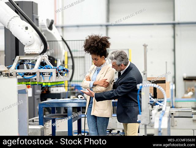 Engineer explaining robotic arm to developer standing with arms crossed in factory
