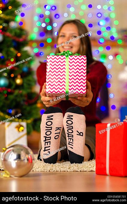 a happy girl in funny socks with the inscription ""who's good? I'm good!"" with a big gift in the background of Christmas lights