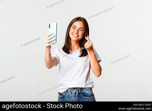 Happy beautiful young woman showing heart gesture and taking selfie on smartphone and smiling