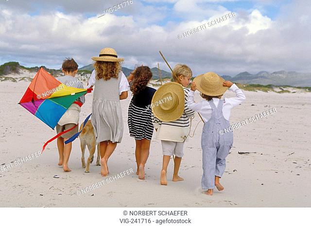 beach-scene, group of 5 children in the age of 7-10 years viewed from the back walking with a dog and a coulered kite, strawhats and sticks bare feeted along...