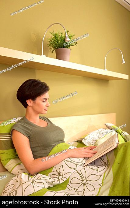 Mid-adult pretty woman reading book, sitting in bed in bedroom