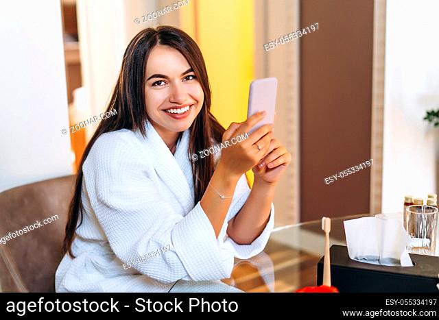 Young woman in bathrobe in hotel room using mobile phone, relaxed after taking a bath