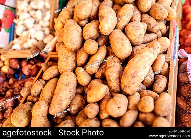 A closeup of potatoes of different shapes and sizes in wooden boxes. There are other vegetables like onions and garlic on the blurred background