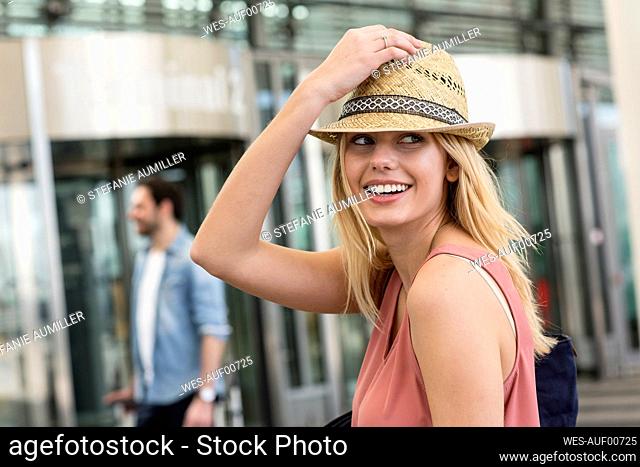 Young woman wearing trilby with boyfriend standing in background outside airport terminal