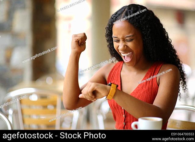 Excited black woman checking smartwatch siting in a bar terrace