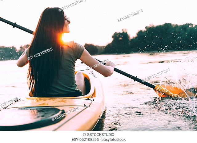 Enjoying perfect sunset on river. Rear view of beautiful young woman kayaking on river and with sunset in the background