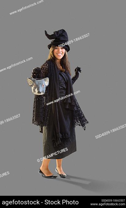 Full length shot of a Hispanic woman dressed as a witch for Halloween holding a mouse head trick or treat bucket dancing, against a white background