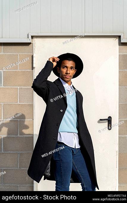 Fashionable young man holding hat while standing against door on sunny day