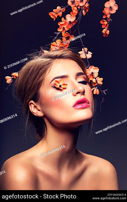 Beautiful young woman with make-up and loose hairdo. Artificial sakura branch with orange flowers hanging over head. Beauty shot on dark background