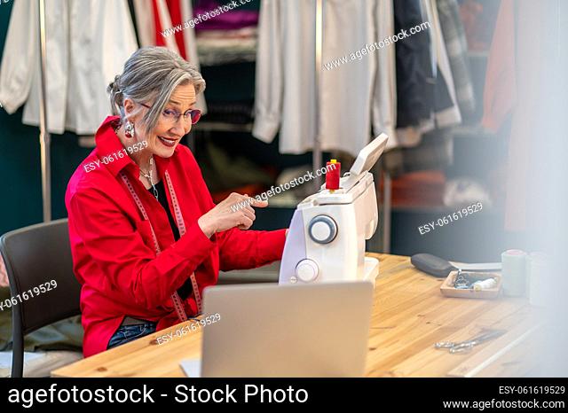 Knowledge. Smiling experienced gray-haired woman in glasses looking at laptop screen pointing at sewing machine sitting at table in atelier