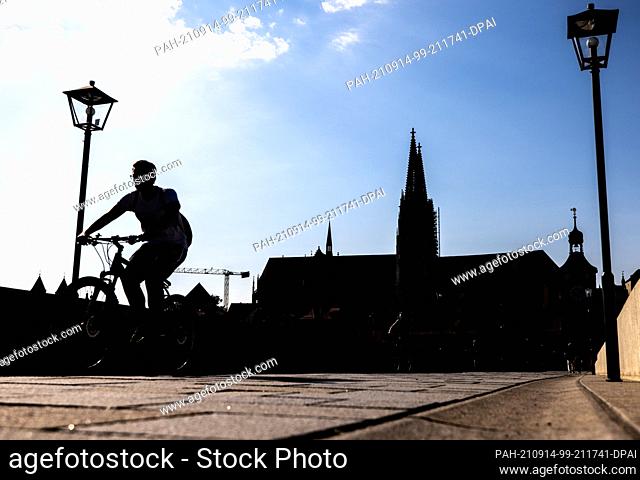 14 September 2021, Bavaria, Regensburg: A cyclist rides over the Steinerne Brücke, while the silhouette of Regensburg Cathedral can be seen in the background