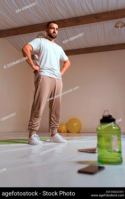 Strong athletic handsome man in white t-shirt and sweatpants taking a break standing at hall in his apartment. Pensive sportsman with thoughtful face ponders...