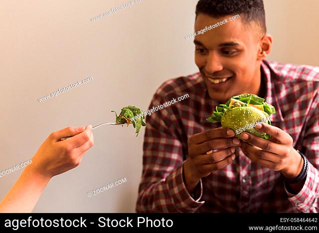 Happy couple spending free time in vegan restaurant or cafe. Portrait of lady eating vegan dish and feeding her boy-friend