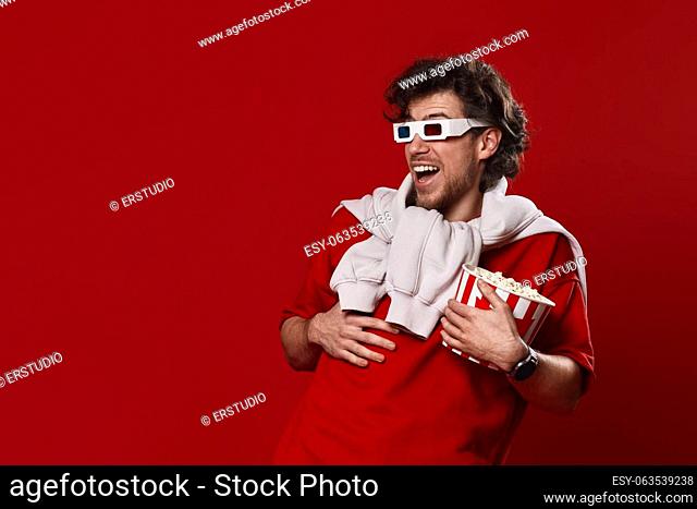 laughing bearded man in 3d-glasses, t-shirt with big bucket of popcorns on red background