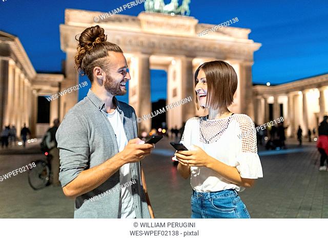 Happy couple using smartphones at Brandenburg gate at blue hour, Berlin, Germany
