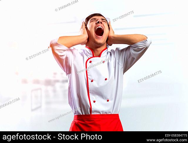 Young male chef screaming in shock and horror. Emotional caucasian chef in white hat and red apron standing with open mouth in light kitchen interior