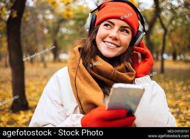 Smiling young woman holding mobile phone listening to music through headphones at park